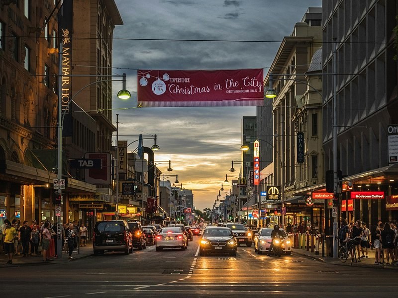 A city-centre street at dusk, shopfronts lit and a Christmas banner overhead